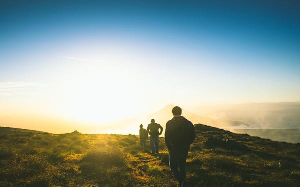 A few travelers walk in a line toward the sunrise and toward the top of a hill